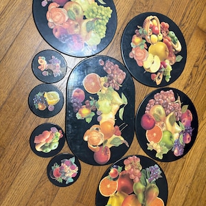 May include: A collection of black serving trays and coasters with vibrant fruit illustrations. The trays are round and oval, featuring apples, grapes, pears, and oranges. The coasters have similar fruit designs. A decorative set for serving food and drinks.