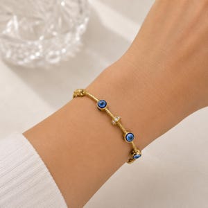 May include: A gold-toned chain bracelet featuring multiple circular blue and white evil eye charms interspersed with a small, cylindrical bead encrusted with clear, sparkling stones. The delicate metal chain has a textured, woven appearance.
