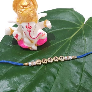May include: A blue string bracelet spelling "ARYAN" in gold letter beads, with pearl and rhinestone accents. A small white and gold deity statue rests above the bracelet on a green leaf, a traditional Indian design.