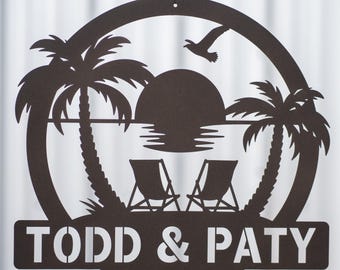 Puede incluir: Una pieza de arte mural de metal marrón que representa una escena de playa tropical. El diseño incluye palmeras, un sol poniente, dos sillas de playa y un pájaro volando. El texto "TODD & PATY" se muestra en la parte inferior.