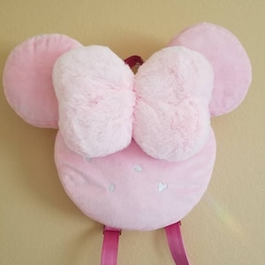 May include: A pink plush backpack shaped like a mouse head with large ears and a fluffy bow. The backpack has pink straps and small white heart-shaped details on the front. The overall design is soft and whimsical.