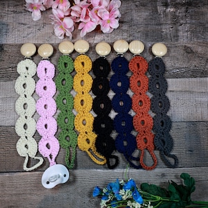 May include: A collection of colorful crochet pacifier clips in various colors, including cream, pink, green, yellow, black, navy, orange, and gray. Each clip features a wooden button and a loop for attaching a pacifier. The clips are arranged on a wooden surface.