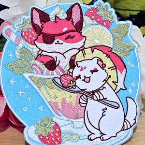 Könnte beinhalten: Ein gestickter Aufnäher mit einem roten Fuchs, der ein Augenpflaster trägt, und einer weißen Katze, die einen Weihnachtsmann-Hut trägt. Beide halten Löffel und sind von Erdbeeren, Zitronen und Minzblättern umgeben. Der Aufnäher befindet sich auf einem blauen Hintergrund mit weißen Sternen.