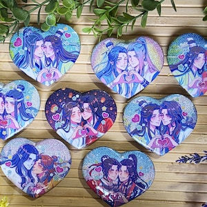 May include: A collection of eight heart-shaped pins featuring anime-style illustrations of pairs of characters with long hair in traditional clothing. Each pin displays a glittery finish and shows the characters forming a heart shape with their hands.
