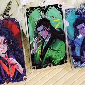 Puede incluir: Tres llaveros acrílicos con ilustraciones de estilo anime de personajes masculinos. Cada llavero tiene un diseño de personaje diferente con esquemas de color únicos, incluyendo rojo, verde y azul. Los llaveros tienen bordes decorativos y herrajes plateados.