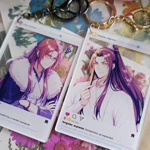 May include: Two rectangular acrylic keychains featuring anime-style illustrations of characters with long purple hair. Each keychain is designed like a social media post with a profile name, caption, and icons for likes, comments, and shares at the bottom.