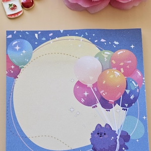 May include: A blue and yellow notepad featuring a purple dog holding colourful balloons. The dog is surrounded by confetti and stars. The notepad has a blank space for writing.
