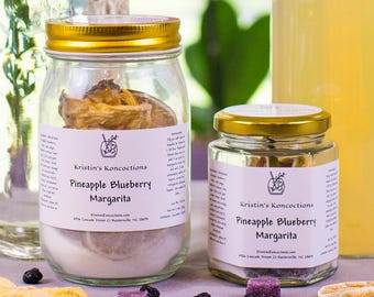 May include: Two glass jars with gold lids and two glass bottles with wooden and black caps. The jars feature labels reading Kristin's Koncoctions Pineapple Blueberry Margarita. Dried pineapple, lime slices, and small purple cubes are scattered nearby.