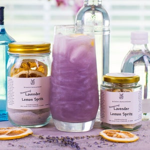 May include: A refreshing lavender-colored cocktail in a tall glass, surrounded by bottles of liquor and jars of ingredients. The jars are labeled "Shimmering Lavender Lemon Spritz" and contain dried lemon slices. The image has a bright, airy feel.
