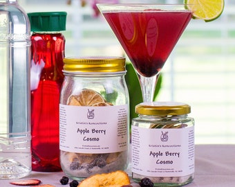 May include: A clear glass bottle with a black cap, a red liquid bottle, two glass jars with gold lids labeled Apple Berry Cosmo, and a martini glass filled with a dark red liquid garnished with a lime slice, with dried fruit slices nearby.