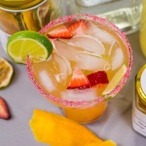 May include: A margarita cocktail with a pink sugar rim, ice, lime wedges, and strawberry slices in a clear glass. The drink is on a gray surface with mango slices and other fruit.