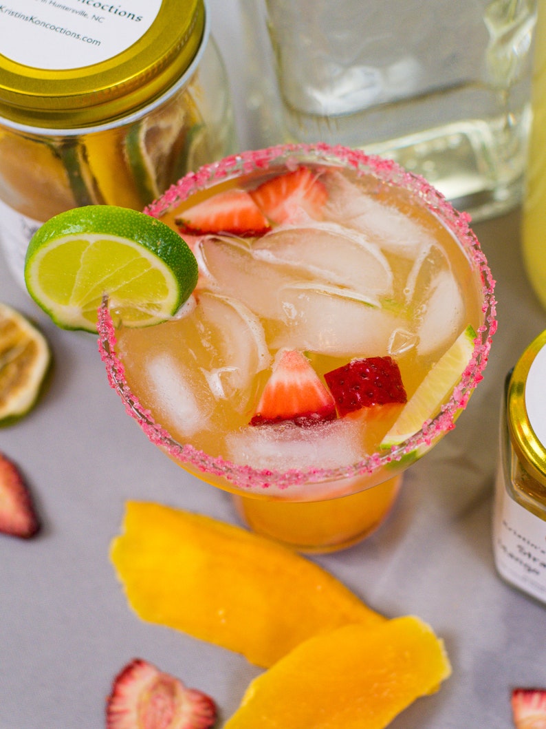 May include: A margarita cocktail with a pink sugar rim, ice, lime wedges, and strawberry slices in a clear glass. The drink is on a gray surface with mango slices and other fruit.