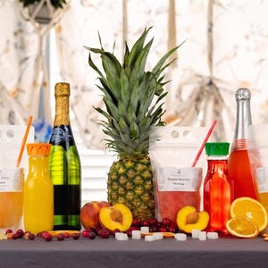 May include: A still life arrangement featuring a pineapple, champagne bottles, juice bottles, and pouches with straws. Sliced oranges, peaches, and a mango are also present. The scene is set on a gray surface with decorative elements.