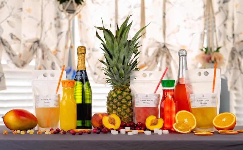May include: A still life arrangement featuring a pineapple, champagne bottles, juice bottles, and pouches with straws. Sliced oranges, peaches, and a mango are also present. The scene is set on a gray surface with decorative elements.