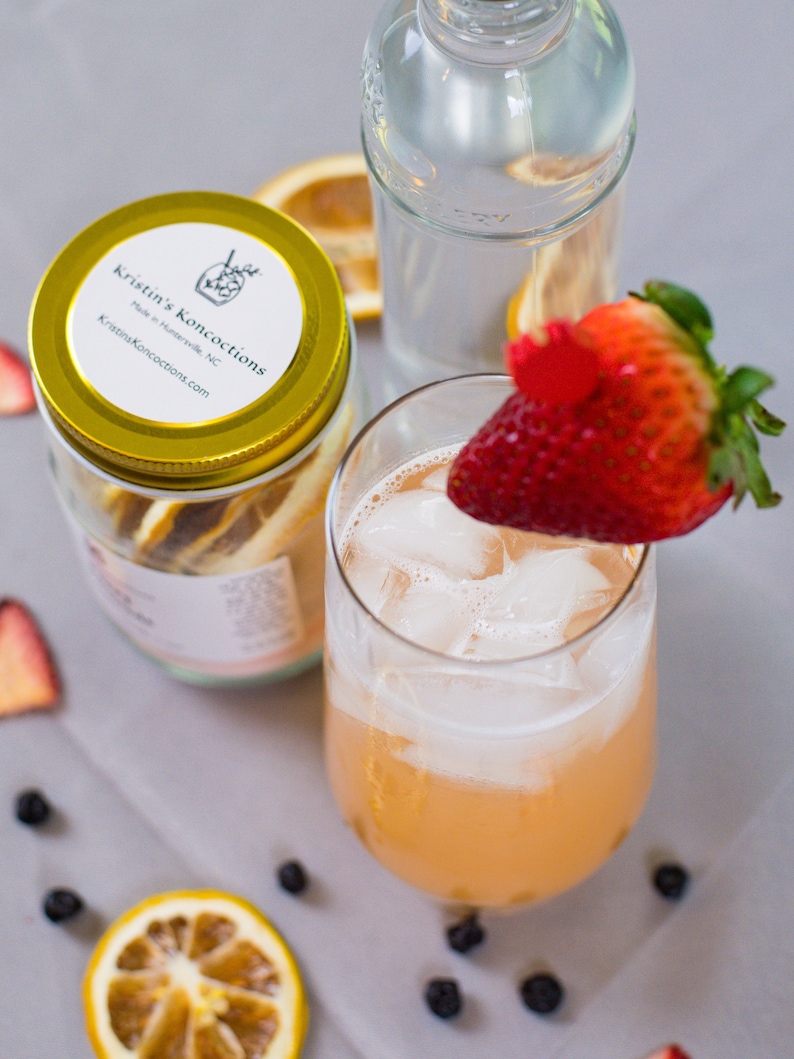 May include: A glass of pink lemonade with ice and a strawberry garnish. The drink is in a clear glass and is surrounded by blueberries and orange slices. The glass is sitting on a gray surface.