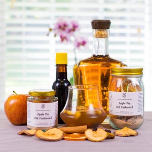 May include: A collection of cocktail ingredients including a large glass bottle of amber liquid, two jars labeled Apple Pie Old Fashioned, a small bottle with a yellow cap, a glass of amber liquid, a fresh apple, and dried fruit slices.
