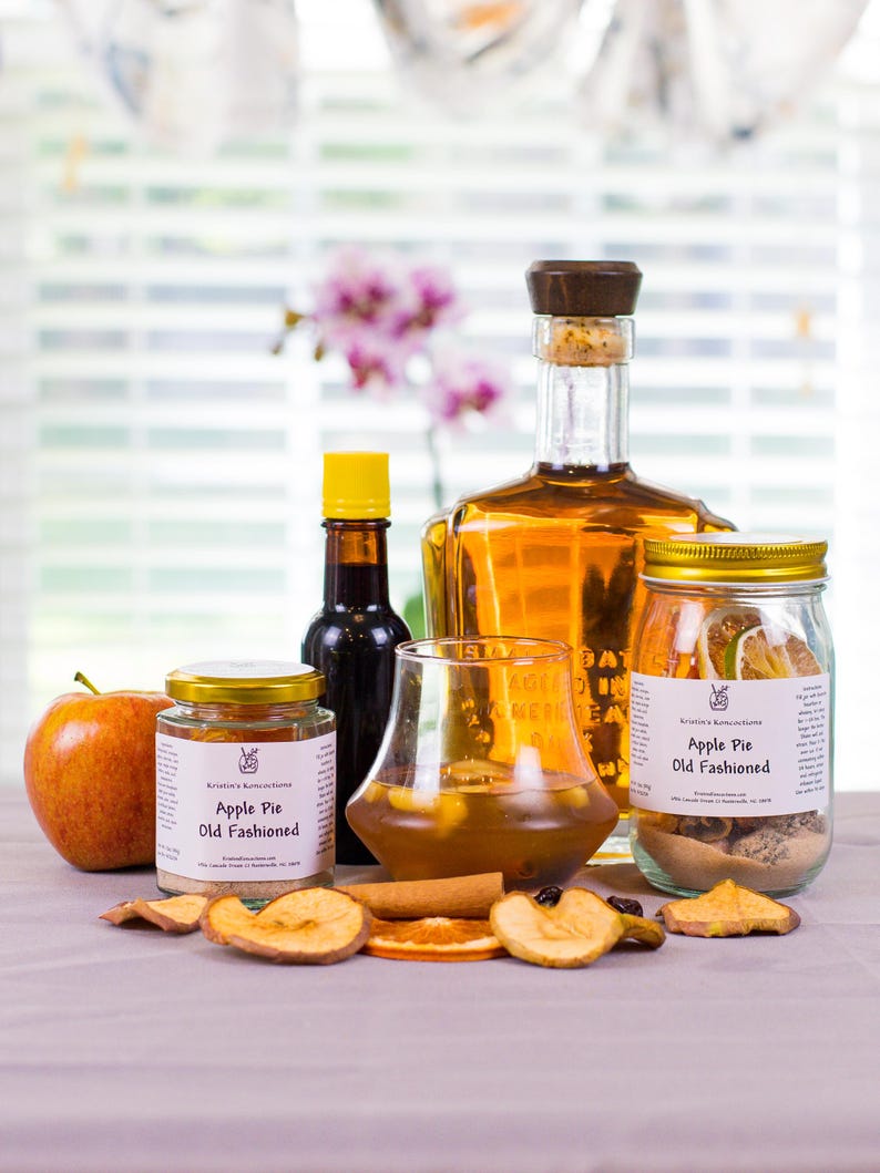 May include: A collection of cocktail ingredients including a large glass bottle of amber liquid, two jars labeled Apple Pie Old Fashioned, a small bottle with a yellow cap, a glass of amber liquid, a fresh apple, and dried fruit slices.