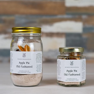 May include: Two clear glass jars with gold lids containing Apple Pie Old Fashioned cocktail infusion mix. Each jar features a white label with the text Kristin's Koncoctions, Apple Pie Old Fashioned, and instructions for use with whiskey.