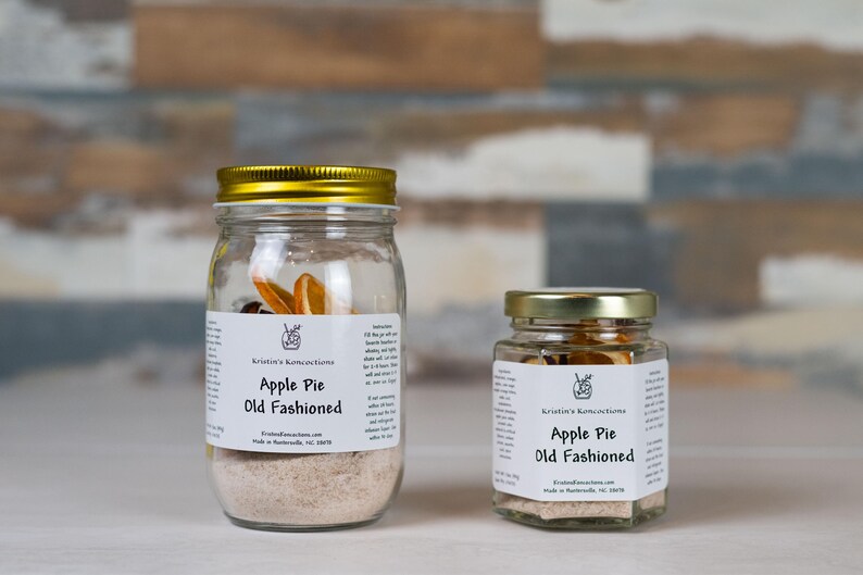 May include: Two clear glass jars with gold lids containing Apple Pie Old Fashioned cocktail infusion mix. Each jar features a white label with the text Kristin's Koncoctions, Apple Pie Old Fashioned, and instructions for use with whiskey.