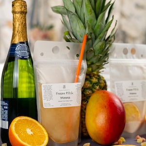 May include: A still life arrangement featuring a bottle of champagne, a pineapple, a mango, and an orange half. A clear pouch labeled "Frozen P.O.G. Mimosa" with a straw is in the center. Orange slices and candied ginger pieces are scattered around the fruit.