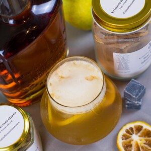May include: A glass of amber-colored cocktail with foam, a bottle of dark liquid, two jars labeled Kristin's Koncoctions, a fresh green pear, two dark gray cubes, and dried citrus slices. The labels feature the text Kristin's Koncoctions.