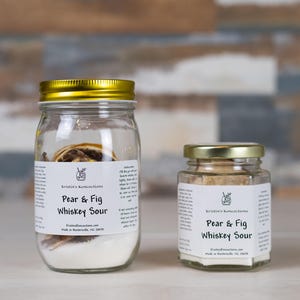 May include: Two clear glass jars filled with Pear & Fig Whiskey Sour cocktail infusion mix. The larger jar has a gold lid, and the smaller jar has a silver lid. Both feature white labels with black text reading Kristin's Rumcoctions.