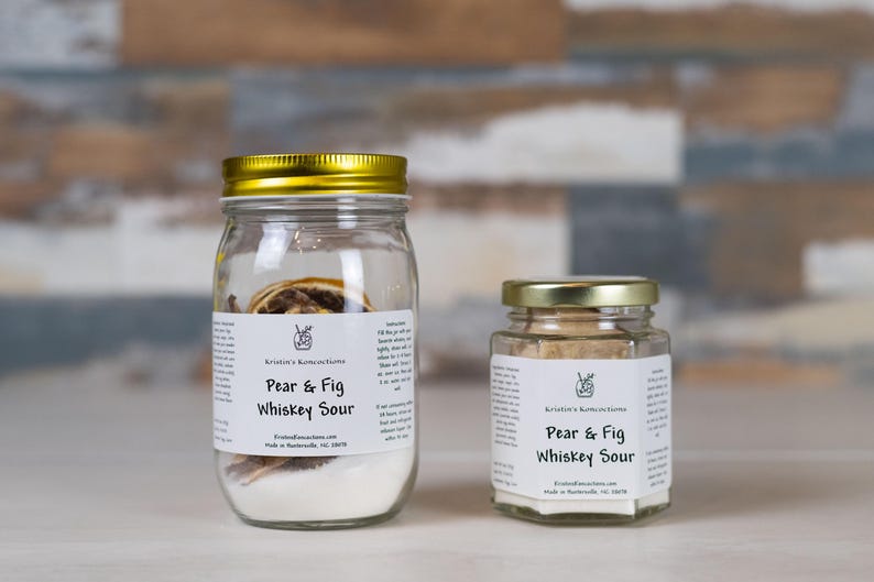 May include: Two clear glass jars filled with Pear & Fig Whiskey Sour cocktail infusion mix. The larger jar has a gold lid, and the smaller jar has a silver lid. Both feature white labels with black text reading Kristin's Rumcoctions.