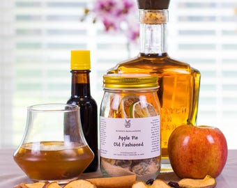 May include: A glass jar labeled Kristin's Koncoctions Apple Pie Old Fashioned containing dried fruit and spices, next to a glass of amber liquid, a dark bottle with a yellow cap, a large glass bottle of amber spirit, and a fresh red apple.