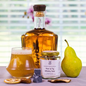 May include: A glass bottle of amber liquid with a wooden stopper, a glass of whiskey sour, a jar labeled Kristin's Koncoctions Pear & Fig Whiskey Sour, a fresh green pear, two dark grey ice cubes, a dried orange slice, and dried fruit pieces.