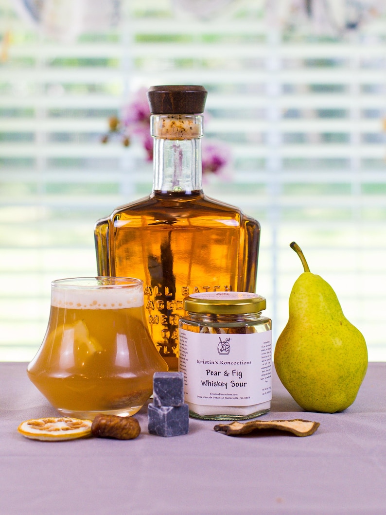 May include: A glass bottle of amber liquid with a wooden stopper, a glass of whiskey sour, a jar labeled Kristin's Koncoctions Pear & Fig Whiskey Sour, a fresh green pear, two dark grey ice cubes, a dried orange slice, and dried fruit pieces.