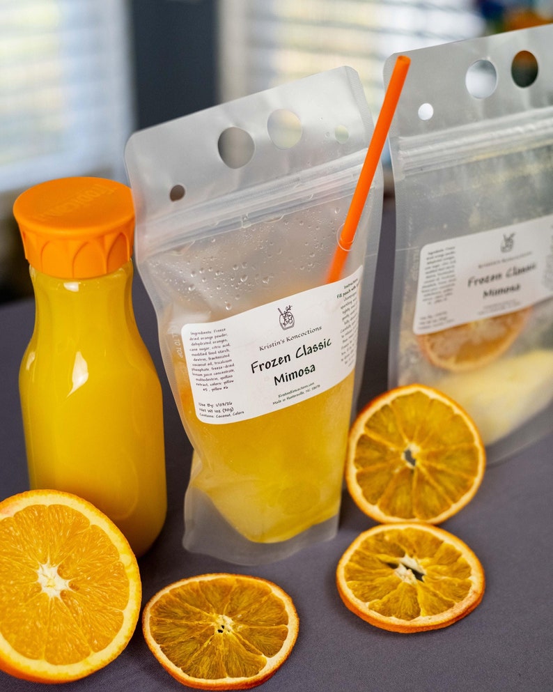 May include: A bottle of orange juice with an orange cap and a clear pouch of frozen mimosa with an orange straw. The pouch is labeled "Frozen Classic Mimosa" with the brand name "Kristin's Konfections". Sliced dried oranges are arranged around the drinks.