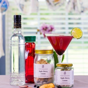 May include: A pink cocktail in a martini glass with a lime wedge garnish. Two glass jars with gold lids contain dried fruit and the text "Apple Berry Cosmo" on the label. A clear glass bottle with a black cap and a red bottle with a green cap are also in the image.