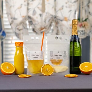 May include: A mimosa kit featuring a bottle of champagne, a bottle of orange juice, and two pouches labeled "Frozen Classic Mimosa." The kit includes orange slices for garnish. The background has floral curtains.