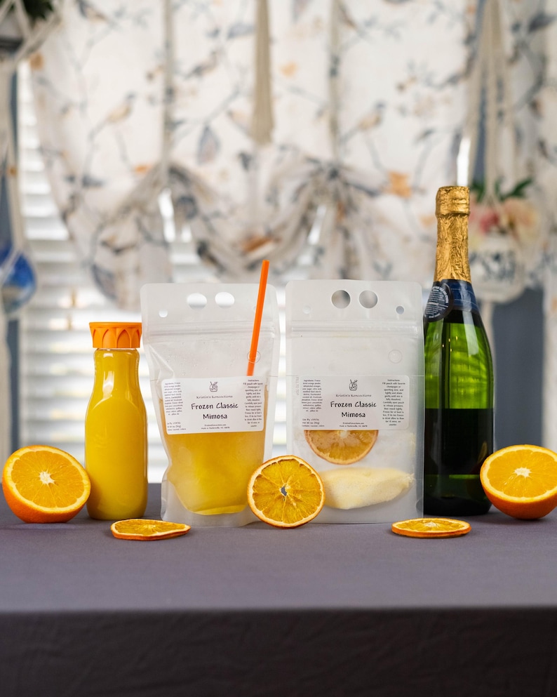 May include: A mimosa kit featuring a bottle of champagne, a bottle of orange juice, and two pouches labeled "Frozen Classic Mimosa." The kit includes orange slices for garnish. The background has floral curtains.