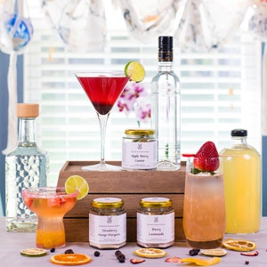 May include: A variety of cocktail mixers in glass jars with gold lids. The jars are labeled "Strawberry Mango Margarita", "Berry Lemonade", and "Apple Berry Cosmo". There are also four cocktails in glasses, including a red cosmo, a pink lemonade, a margarita, and a clear drink with a strawberry garnish.