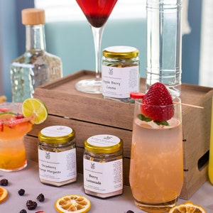 May include: A set of four cocktail mix jars with gold lids and labels. The jars are arranged on a wooden surface with a glass of red cocktail, a glass of pink lemonade, and a bottle of vodka. The labels on the jars read "Apple Berry Cosmo", "Strawberry Mango Margarita", "Berry Lemonade", and "Cranberry & Lime Vodka".
