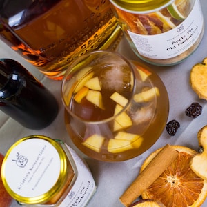 May include: A glass of amber-colored cocktail with apple chunks and ice, surrounded by two jars labeled Kristin's Koncoctions Apple Pie Old Fashioned, a bottle of dark liquid, a cinnamon stick, dried orange slices, and dried apple slices.