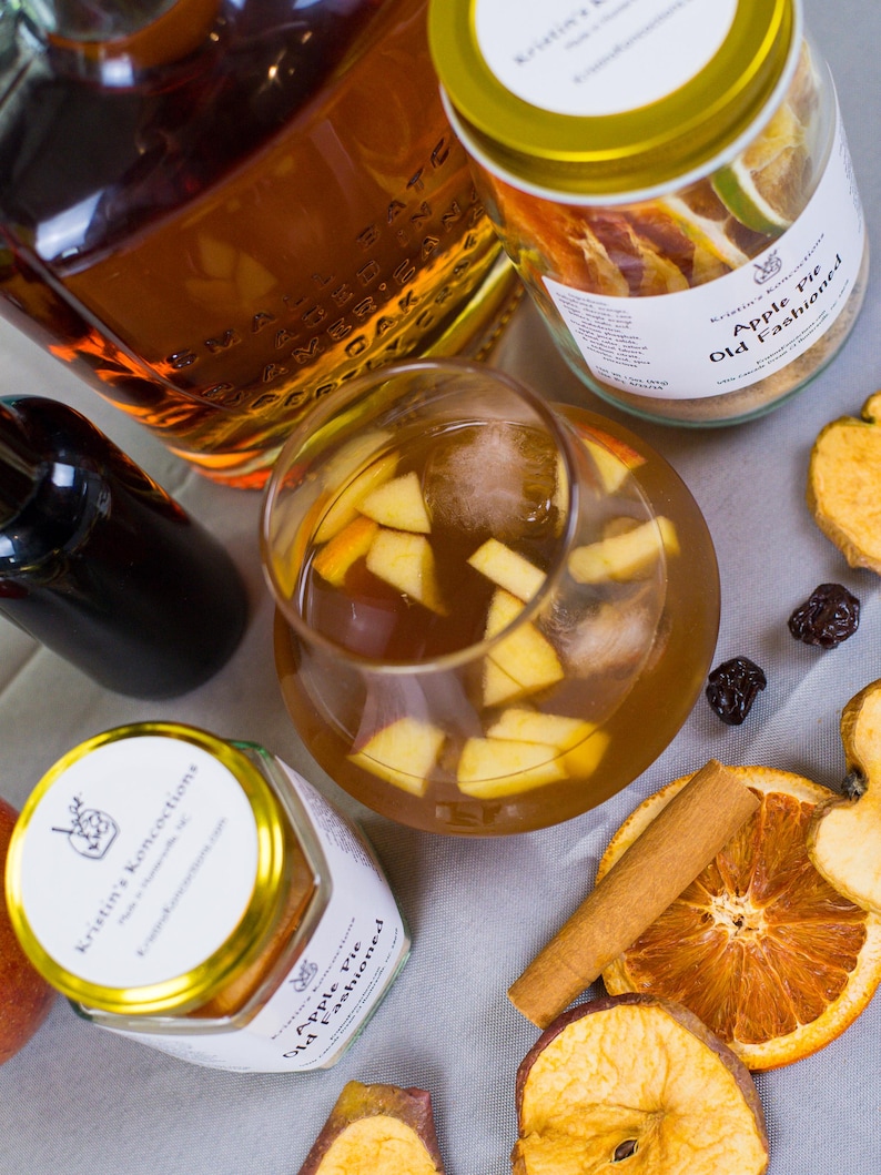 May include: A glass of amber-colored cocktail with apple chunks and ice, surrounded by two jars labeled Kristin's Koncoctions Apple Pie Old Fashioned, a bottle of dark liquid, a cinnamon stick, dried orange slices, and dried apple slices.