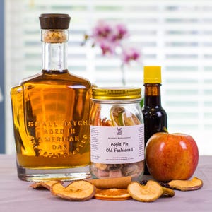 May include: A glass bottle of amber liquid with a wooden stopper, a jar labeled Apple Pie Old Fashioned containing dried fruit, a small dark bottle with a yellow cap, and a fresh red apple surrounded by scattered dried apple and orange slices.