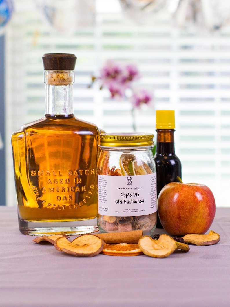 May include: A glass bottle of amber liquid with a wooden stopper, a jar labeled Apple Pie Old Fashioned containing dried fruit, a small dark bottle with a yellow cap, and a fresh red apple surrounded by scattered dried apple and orange slices.