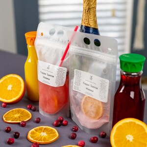May include: A still life arrangement featuring a champagne bottle, juice bottles, and pouches labeled "Frozen Sunrise Mimosa." Garnishes include orange slices and cranberries. The pouches contain a pinkish liquid and a slice of orange.