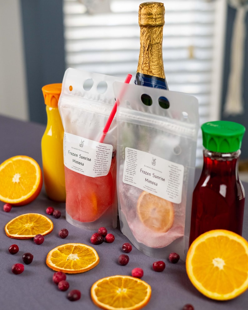 May include: A still life arrangement featuring a champagne bottle, juice bottles, and pouches labeled "Frozen Sunrise Mimosa." Garnishes include orange slices and cranberries. The pouches contain a pinkish liquid and a slice of orange.