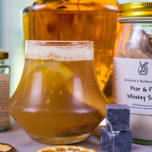 May include: A glass of amber-colored cocktail with a lemon slice, positioned in front of a large amber glass bottle and a jar labeled Kristin's Koncoctions Pear & F Whiskey S. Two dark gray stone ice cubes and a dried citrus slice sit in the foreground.