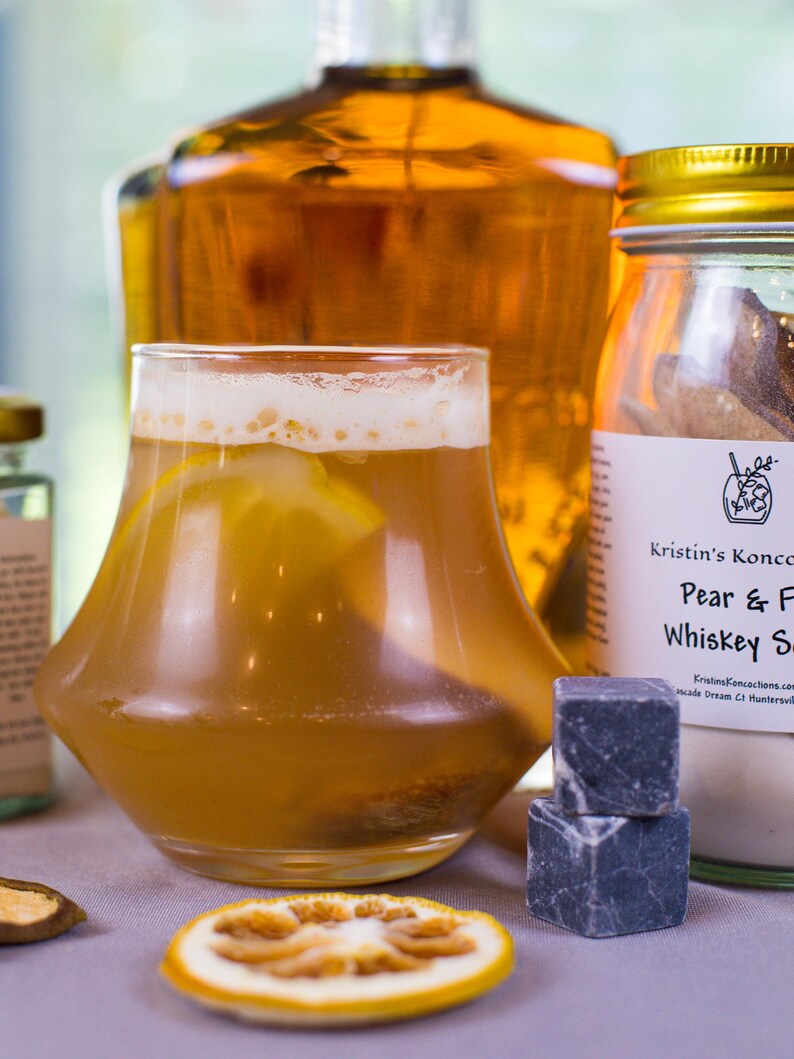 May include: A glass of amber-colored cocktail with a lemon slice, positioned in front of a large amber glass bottle and a jar labeled Kristin's Koncoctions Pear & F Whiskey S. Two dark gray stone ice cubes and a dried citrus slice sit in the foreground.