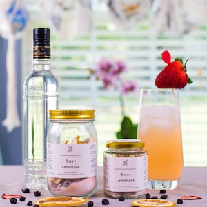 May include: Two glass jars with gold lids filled with dried fruit and a label that reads "Kristin's Konections Berry Lemonade". A glass of pink lemonade with a strawberry garnish sits on a table with dried fruit slices and blueberries.