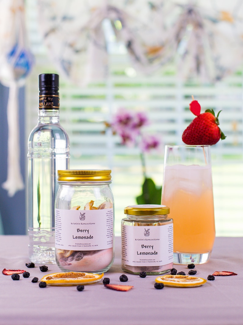 May include: Two glass jars with gold lids filled with dried fruit and a label that reads "Kristin's Konections Berry Lemonade". A glass of pink lemonade with a strawberry garnish sits on a table with dried fruit slices and blueberries.