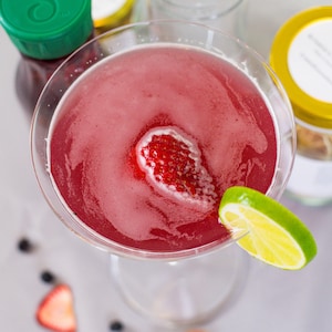 May include: A pink cocktail in a martini glass garnished with a strawberry and a lime wedge. The cocktail is surrounded by other ingredients, including a bottle of juice, a jar of honey, and a bottle of vodka.