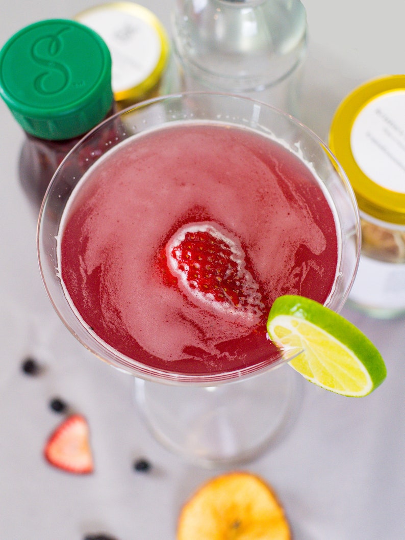 May include: A pink cocktail in a martini glass garnished with a strawberry and a lime wedge. The cocktail is surrounded by other ingredients, including a bottle of juice, a jar of honey, and a bottle of vodka.