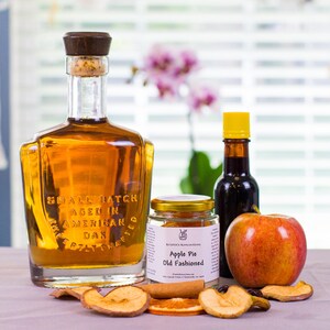 May include: A glass bottle of amber liquid with a dark wood stopper, a small jar labeled Apple Pie Old Fashioned, a dark glass bottle with a yellow cap, a fresh red apple, and dried fruit slices including apple and orange pieces.