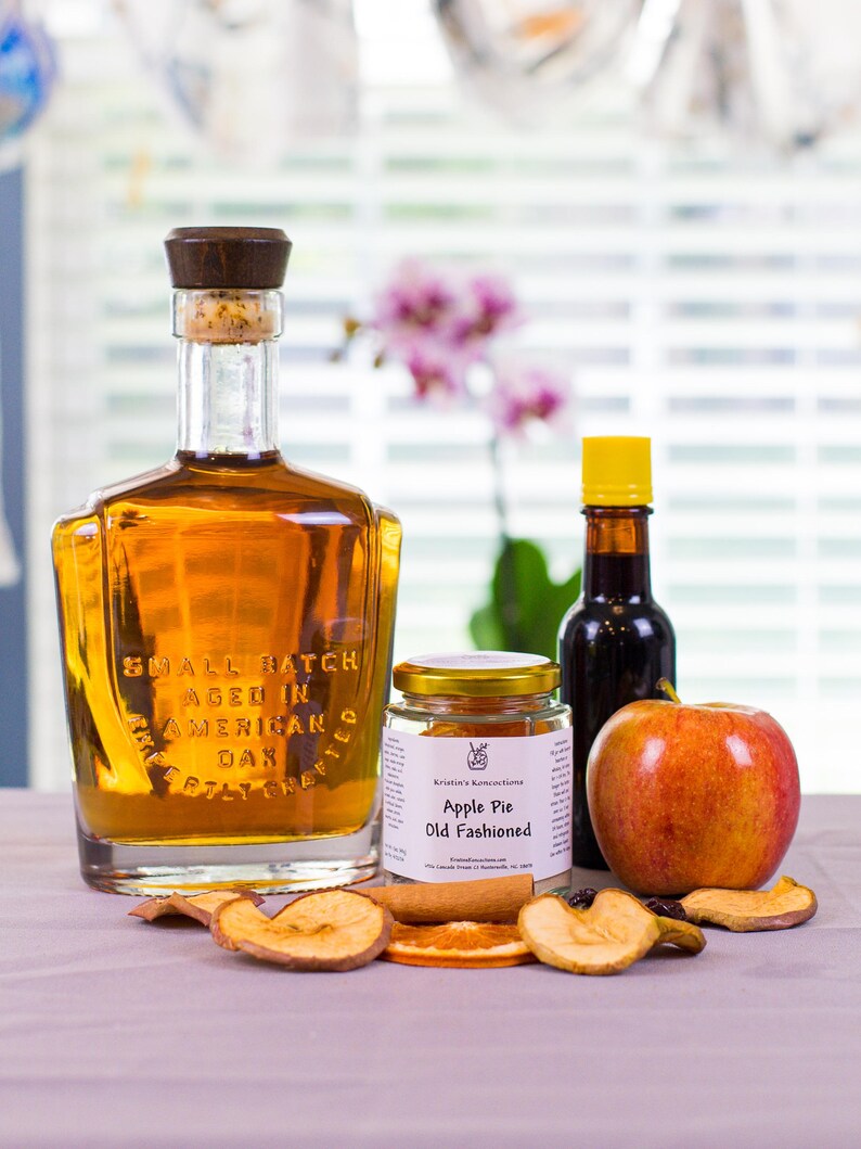 May include: A glass bottle of amber liquid with a dark wood stopper, a small jar labeled Apple Pie Old Fashioned, a dark glass bottle with a yellow cap, a fresh red apple, and dried fruit slices including apple and orange pieces.